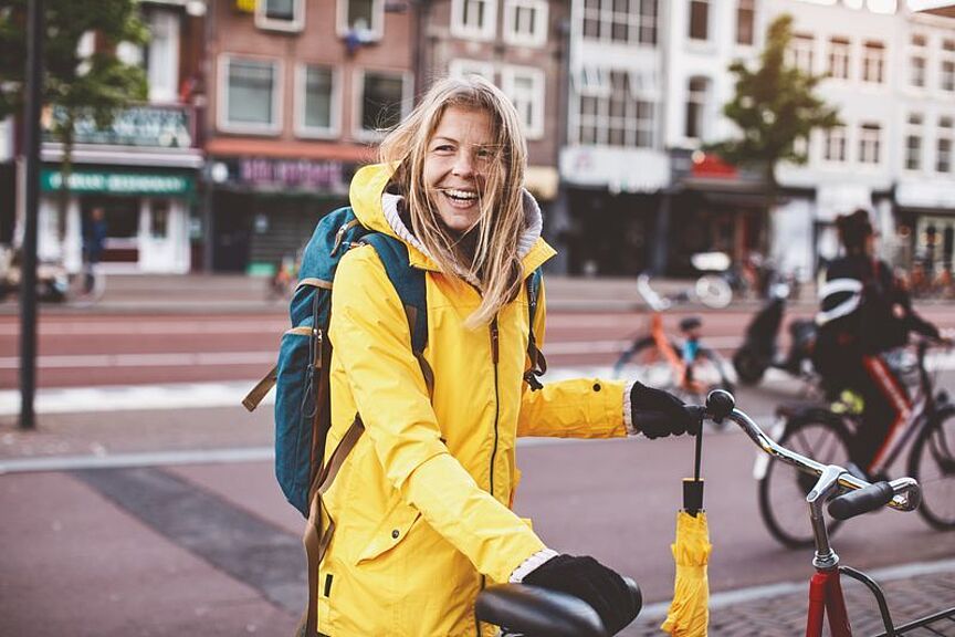 Weltfrauentag Symbolbild: Eine Frau in einer gelben Regenjacke steht lachend an ihrem Fahrrad, bereit loszufahren.